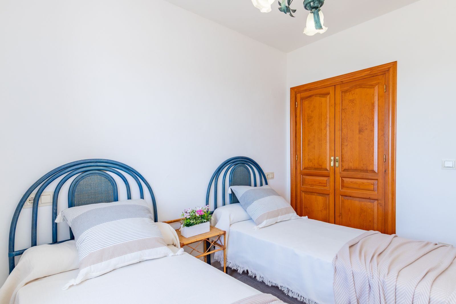 Dormitorio con dos camas individuales, cabeceros de mimbre azul y un amplio armario empotrado de madera natural. Espacio luminoso con paredes blancas y suelos cerámicos.