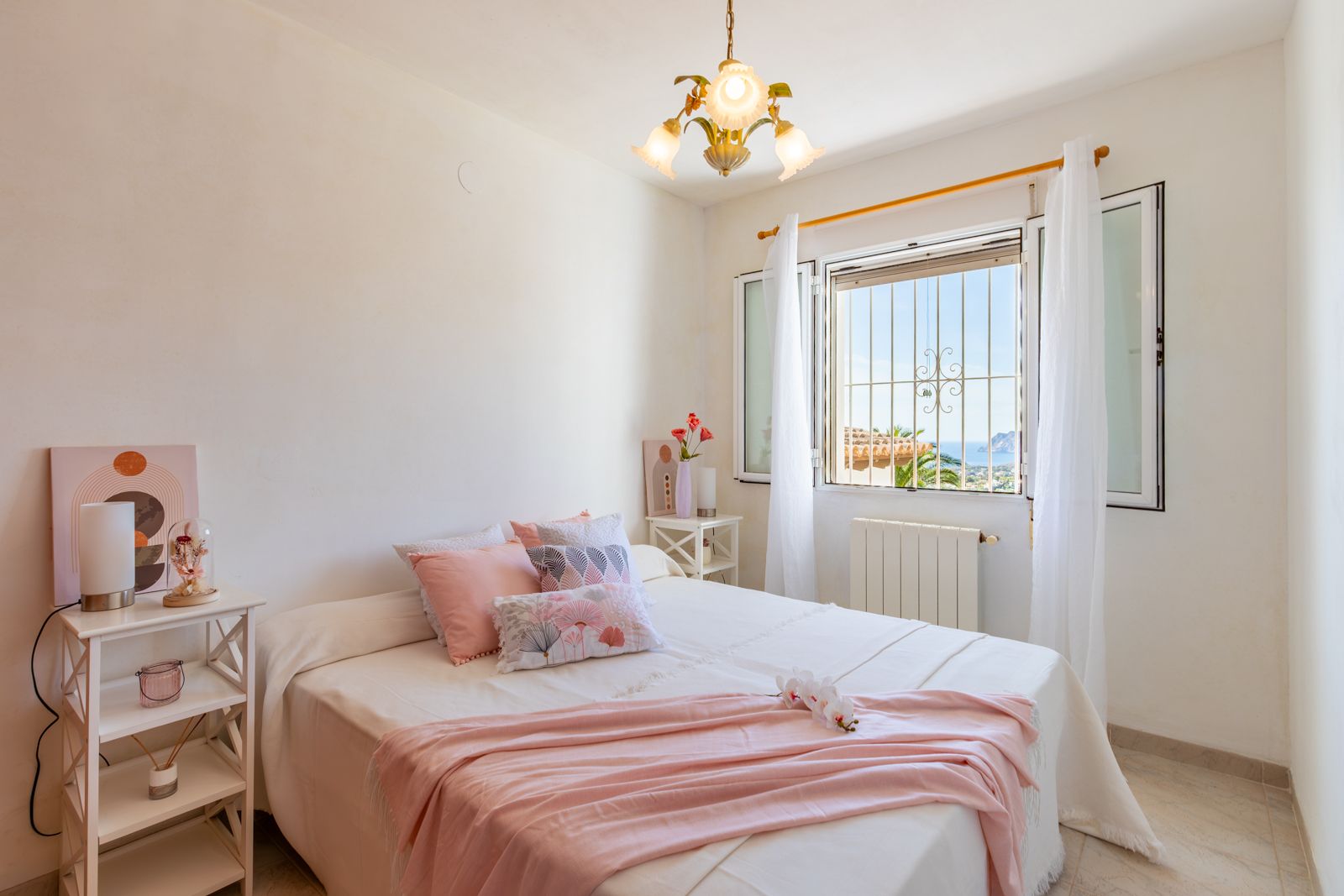 Dormitorio luminoso con cama doble, paredes blancas y suelos cerámicos. La ventana ofrece vistas despejadas al mar y al Peñón de Ifach bajo la luz natural del Mediterráneo.