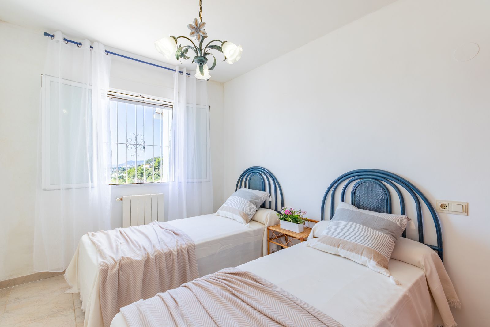 Dormitorio luminoso con dos camas individuales, cabeceros azules de diseño curvo, calefacción central y ventana con vistas despejadas al entorno natural de Moraira.