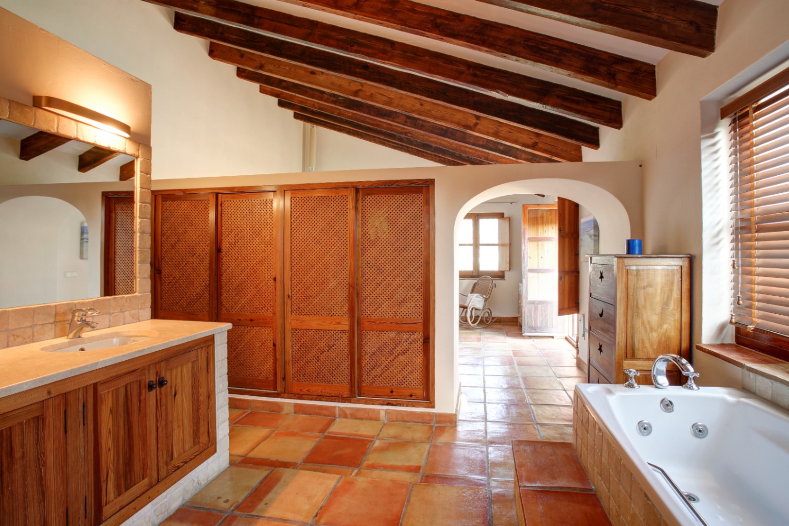 Baño rústico con vigas de madera vista, suelo de barro cocido y bañera de hidromasaje. Incluye armarios de madera con celosía y encimera de mármol crema.