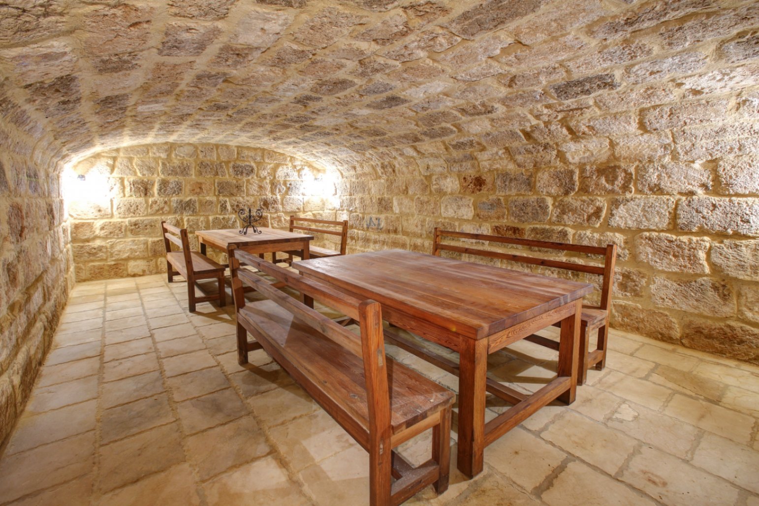 Bodega con techo abovedado de piedra natural, equipada con mesas y bancos de madera maciza sobre un suelo de losas tradicionales y cálida iluminación ambiental.