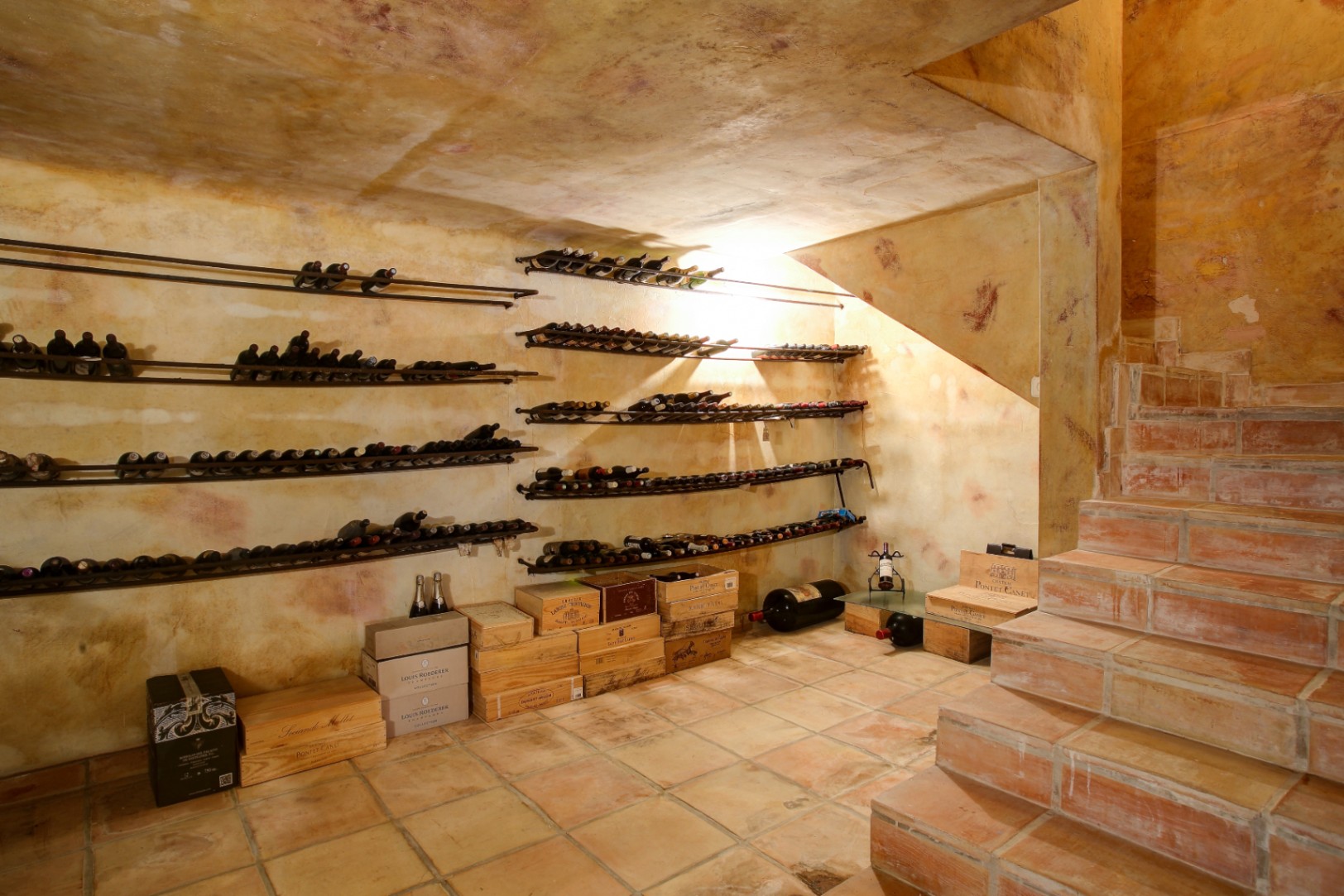 Bodega rústica con suelos de barro cocido, paredes de acabado artesanal y estanterías metálicas de pared para botellas, situada bajo la escalera.