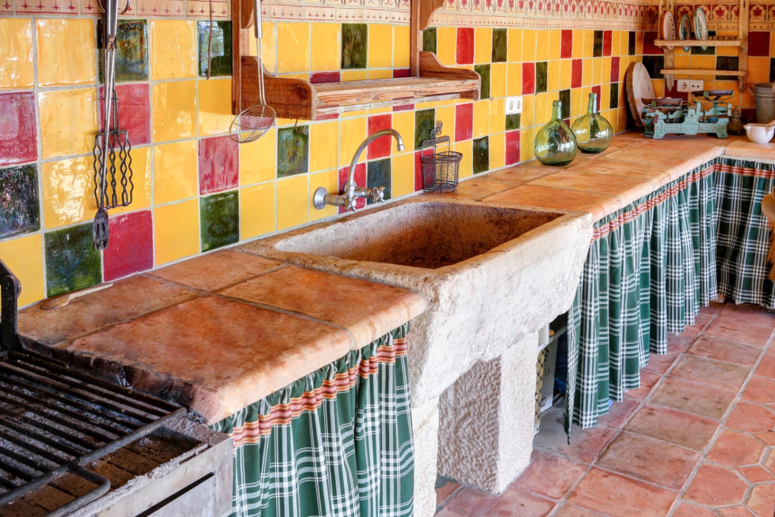 Cocina de verano con fregadero de piedra maciza, encimera de barro cocido y frontal de azulejos artesanales. Incluye zona de barbacoa y almacenaje inferior con cortinas textiles.