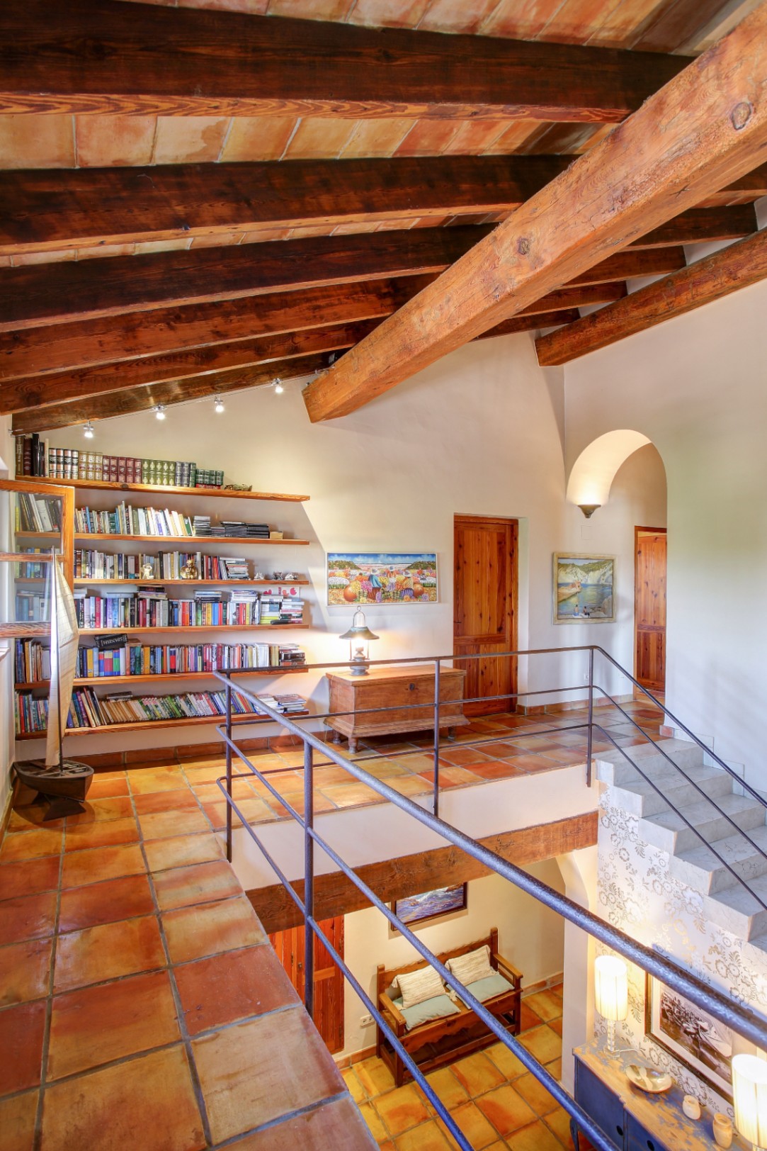 Distribuidor en planta superior con techos de vigas de madera y bovedilla, suelos de barro cocido, biblioteca integrada y vistas al salón de doble altura.