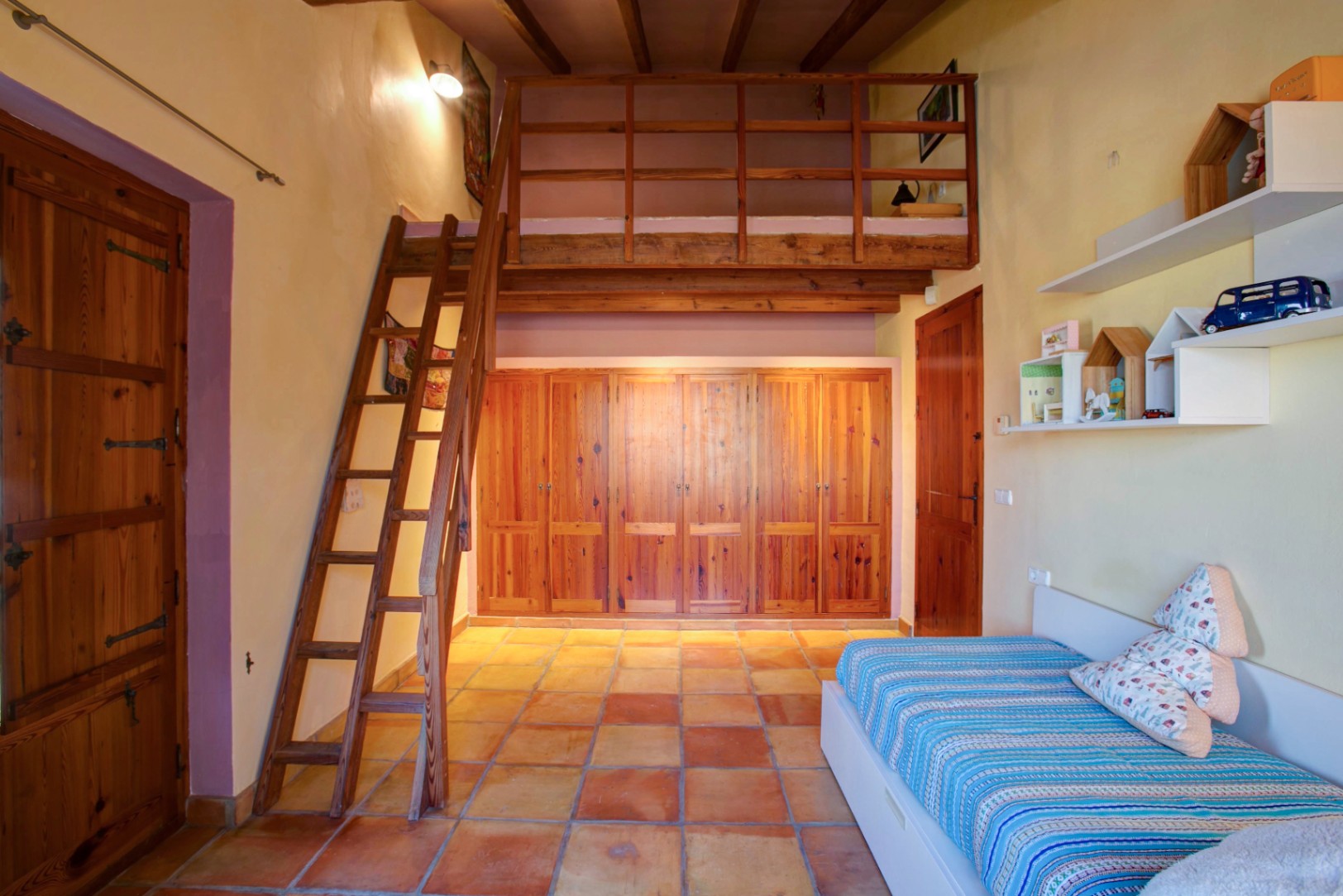 Dormitorio con altillo de madera y escalera, armarios empotrados de madera de mobila y suelo de barro cocido bajo techos de vigas vistas.