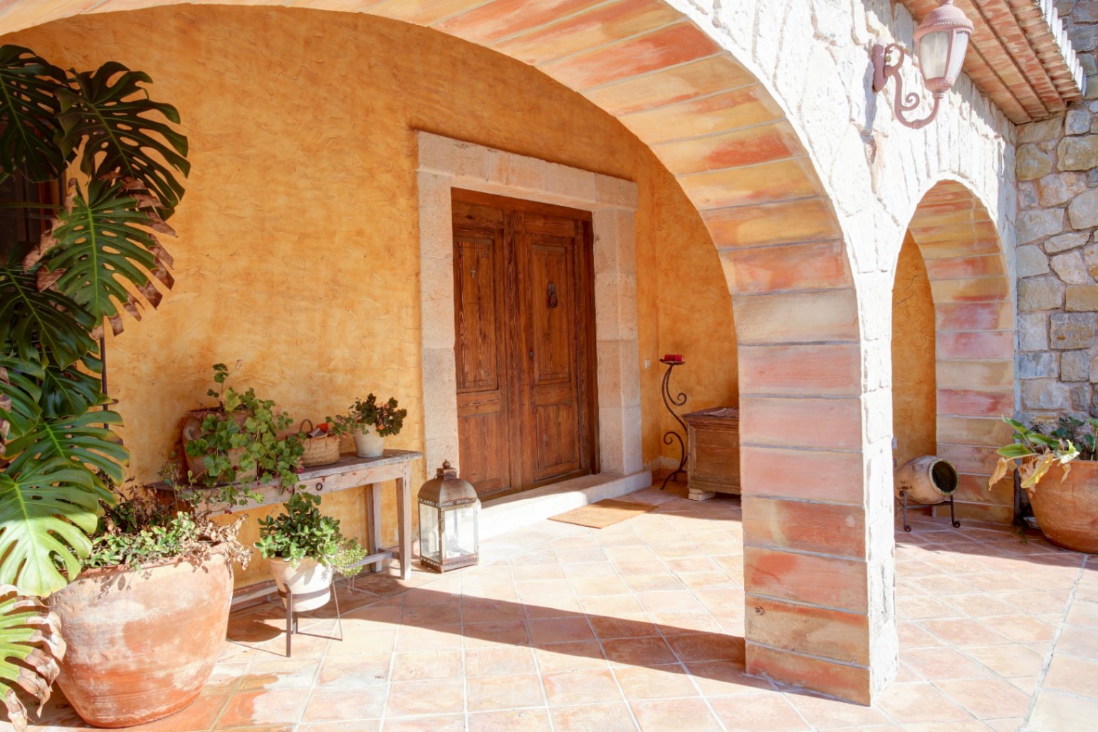 Entrada principal bajo un porche de arcos de piedra y ladrillo, con suelos de barro cocido, puerta de madera maciza y abundante vegetación mediterránea.