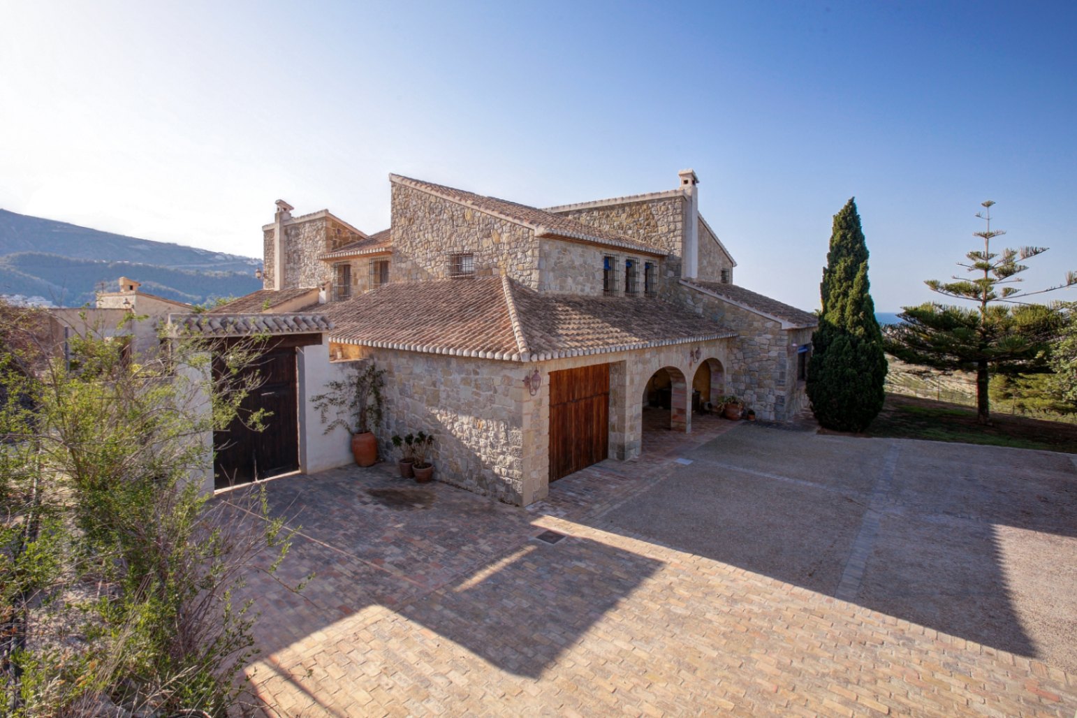 Fachada de piedra natural con arcos tradicionales, tejado de teja cerámica y amplio patio de barro cocido bajo el sol del Mediterráneo.