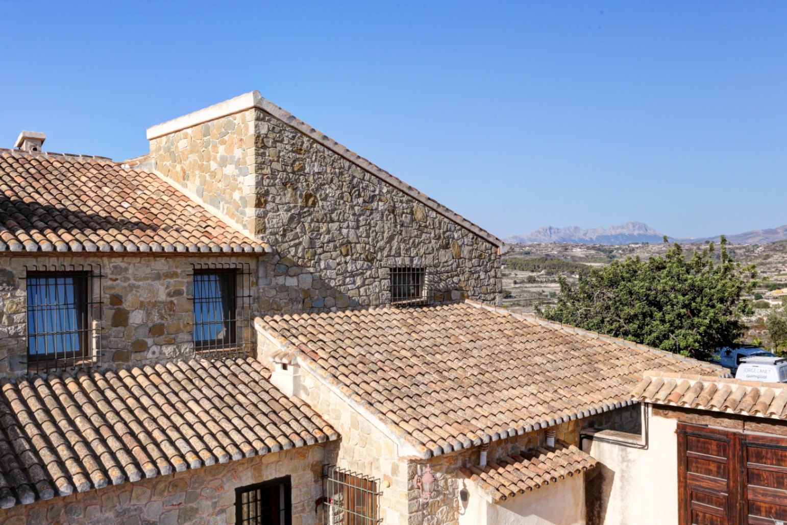 Fachada de piedra natural y cubiertas de teja cerámica con vistas despejadas a las montañas y al entorno rural mediterráneo.