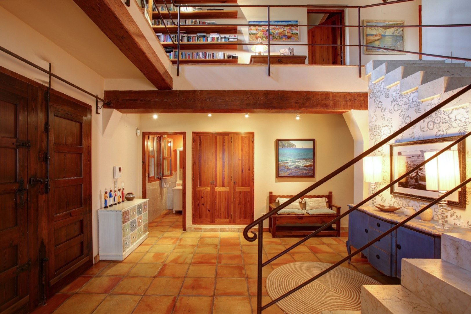 Hall de entrada con suelos de barro cocido, vigas de madera de mobila y una escalera de mármol que conecta con una galería superior destinada a biblioteca.