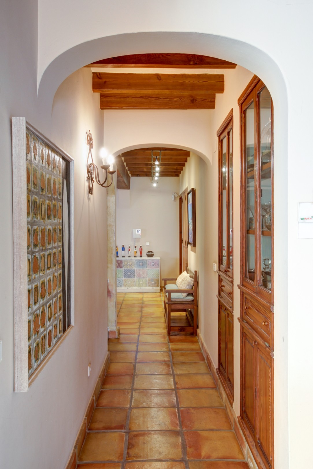 Pasillo con arcos de medio punto, vigas de madera vista y suelo de barro cocido. Incluye vitrinas empotradas de madera noble y una iluminación cálida mediante apliques clásicos.