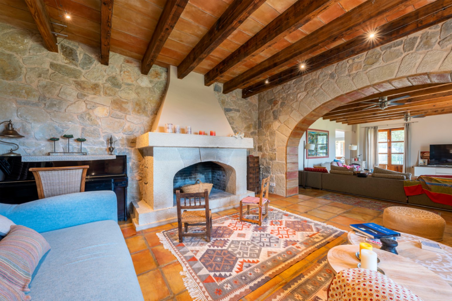 Salón rústico con paredes de piedra natural, chimenea de cantería y techos de vigas de madera. Un gran arco de piedra comunica los ambientes sobre suelos de barro cocido.