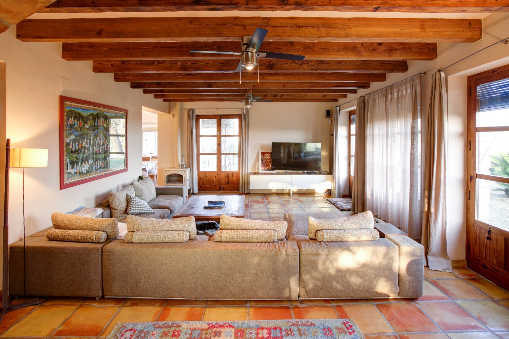 Salón rústico con vigas de madera vista, suelo de barro cocido, una estufa de leña blanca y amplios ventanales de madera que conectan con el exterior.