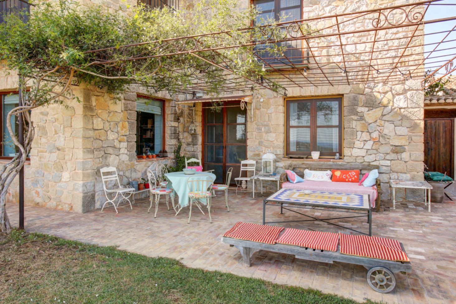 Terraza exterior con fachada de piedra natural, suelo de barro cocido y pérgola de forja con vegetación. Incluye zona de estar con mobiliario rústico y mesa con sobre de mosaico.