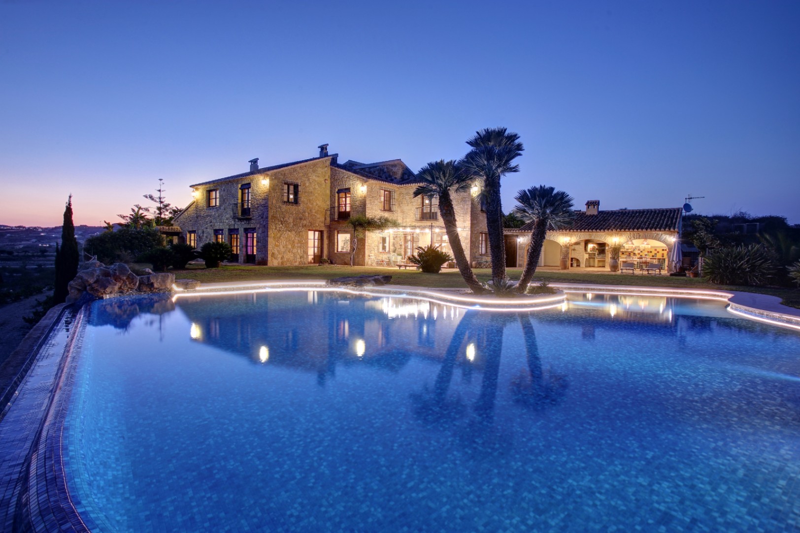 Vista nocturna de la fachada de piedra natural y la piscina de forma orgánica con iluminación LED perimetral, junto a la zona de cocina de verano y barbacoa.