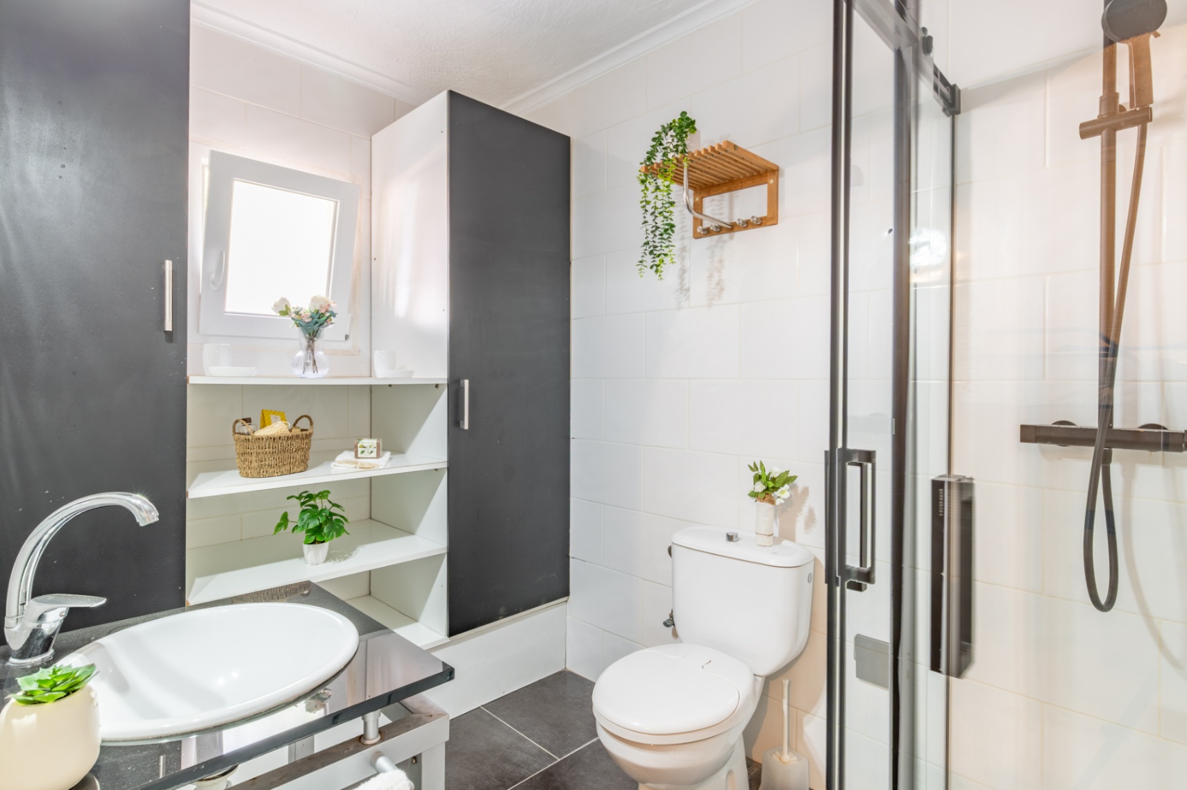 Baño funcional con ducha de mampara de cristal y grifería negra. Incluye lavabo sobre encimera oscura, estanterías integradas y revestimiento cerámico blanco.