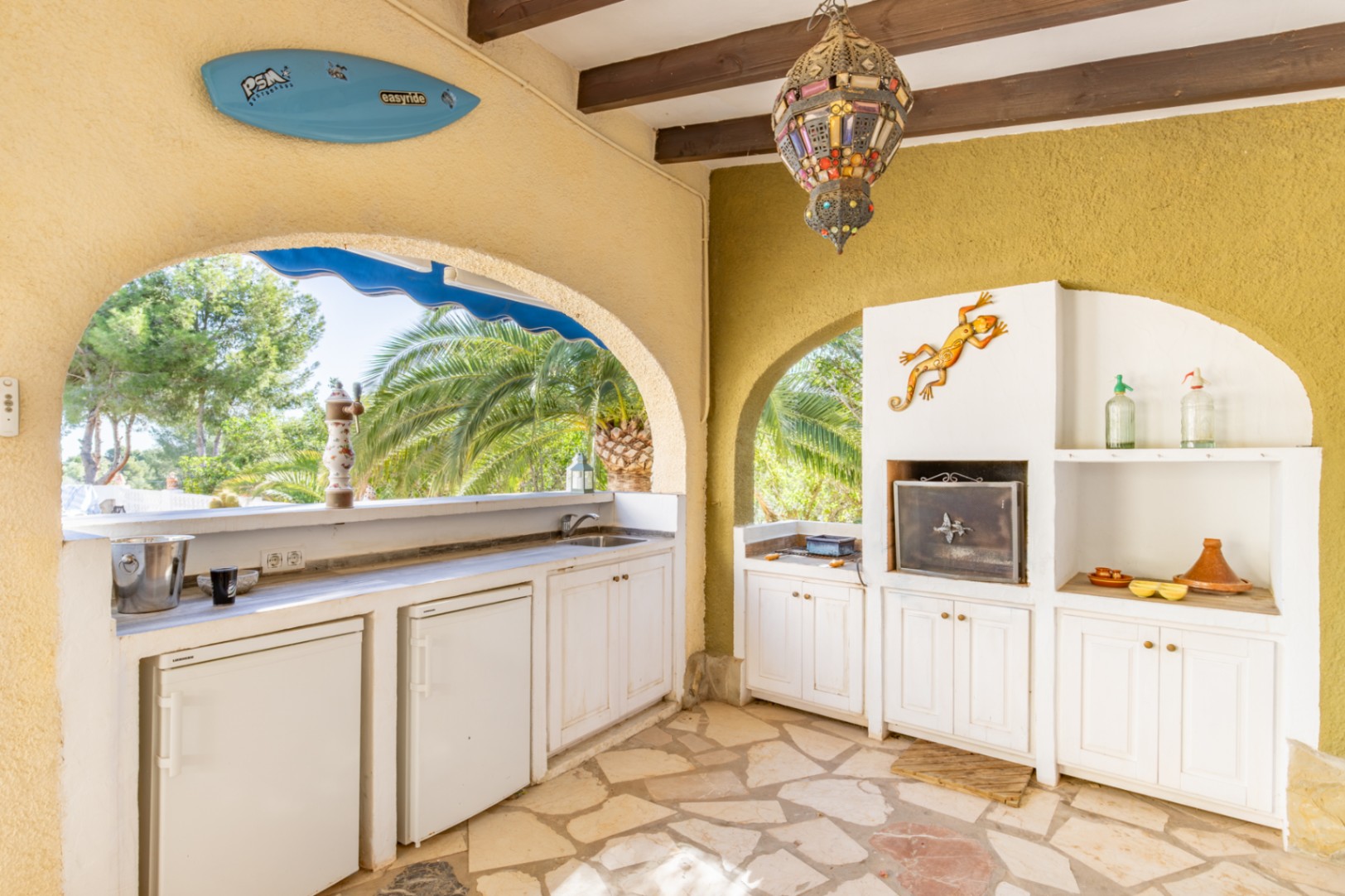 Cocina de verano techada con vigas de madera, equipada con barbacoa de obra, múltiples neveras y fregadero. Cuenta con suelo de piedra natural y vistas al jardín mediterráneo.
