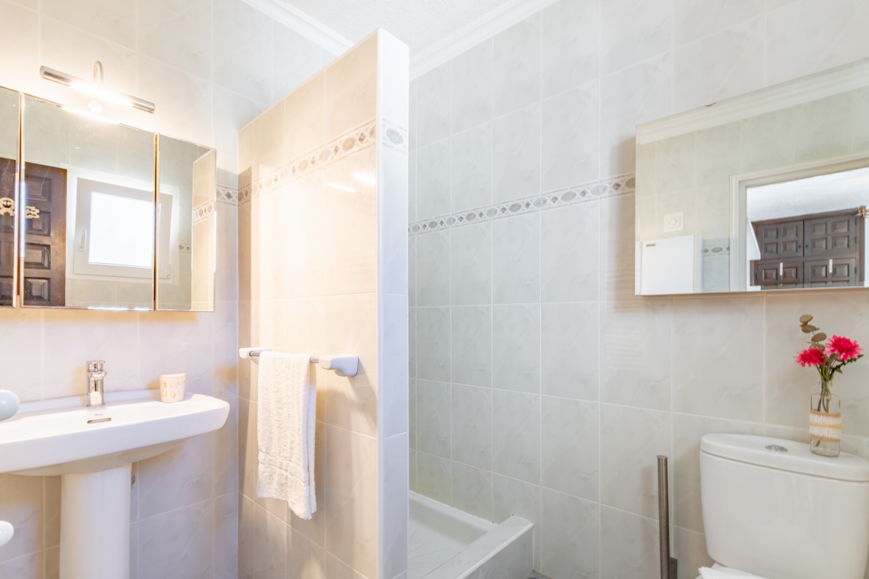 Cuarto de baño con azulejos en tonos crema, lavabo de pedestal, espejo con iluminación superior y zona de ducha separada por un tabique de obra.