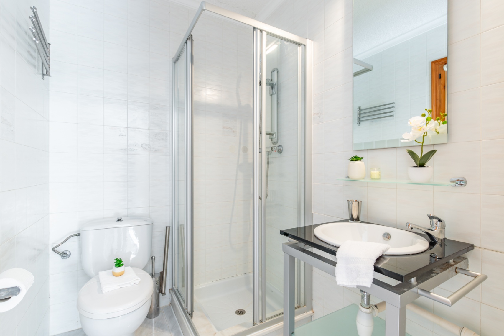 Cuarto de baño funcional con paredes alicatadas en blanco, cabina de ducha con perfilería de aluminio y lavabo encastrado en encimera de piedra oscura.