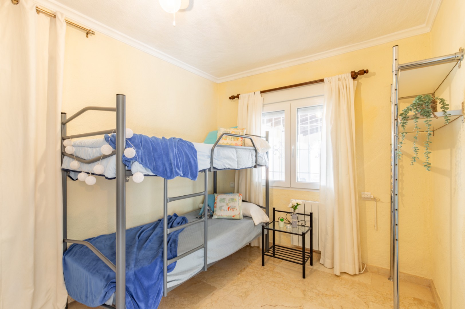 Dormitorio con literas metálicas, paredes en tono crema y suelo de mármol. Incluye ventana con luz natural, cortinas blancas y una estantería abierta lateral.