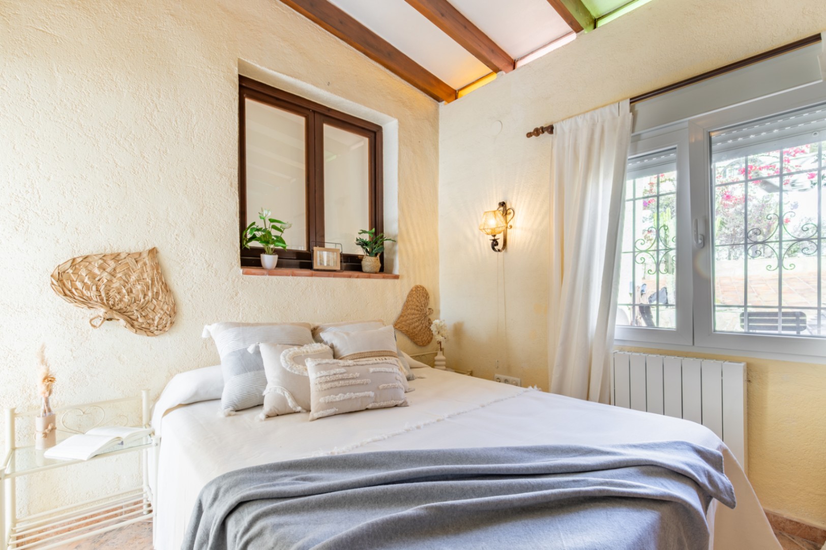 Dormitorio con techos de vigas de madera vistas, paredes de textura rústica y ventanas que aportan luz natural. Incluye radiador y decoración de estilo mediterráneo.