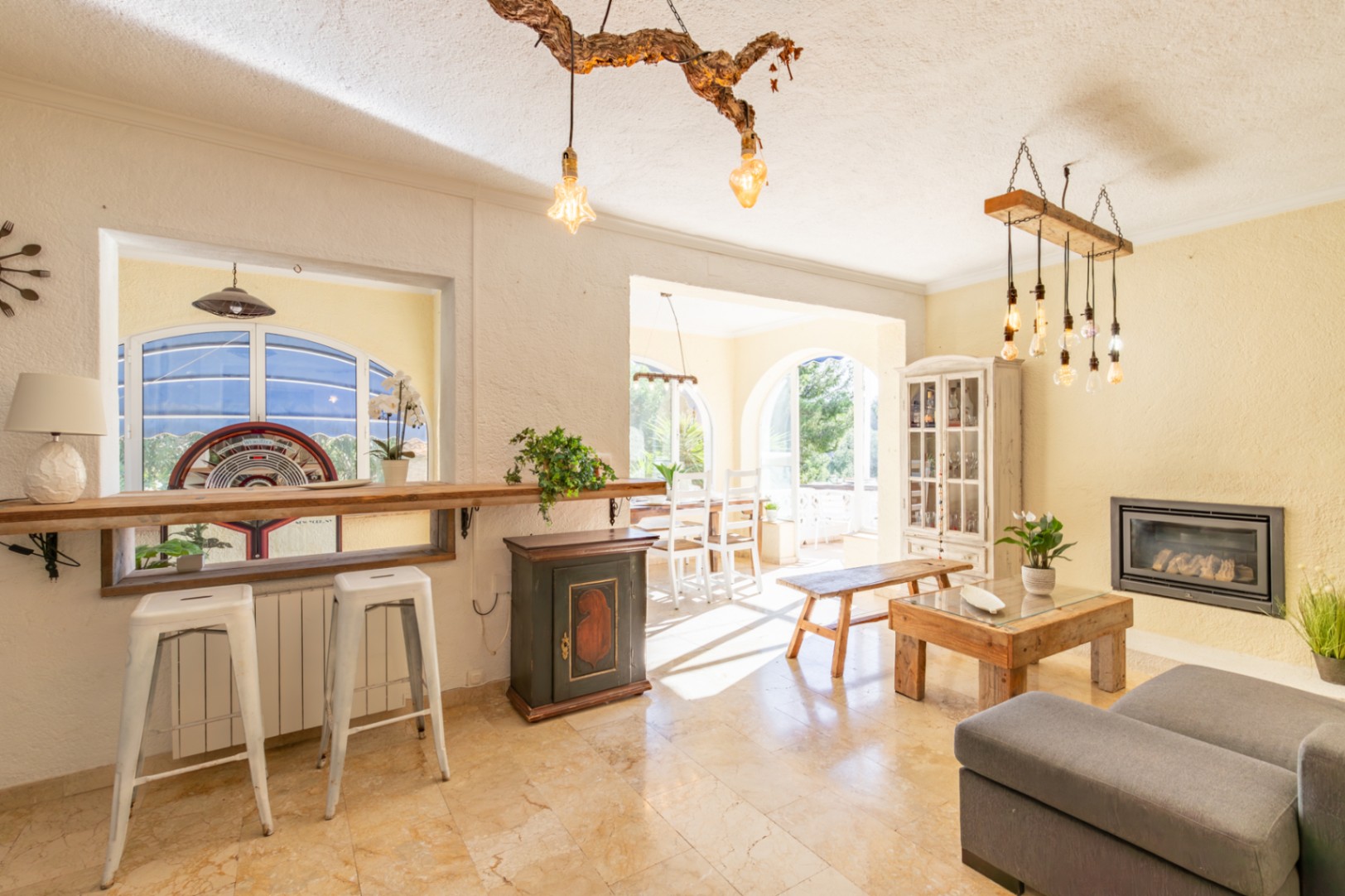 Salón luminoso con suelos de mármol, chimenea encastrada y barra de madera rústica. Destacan las lámparas artesanales y el acceso al comedor mediante arcos.