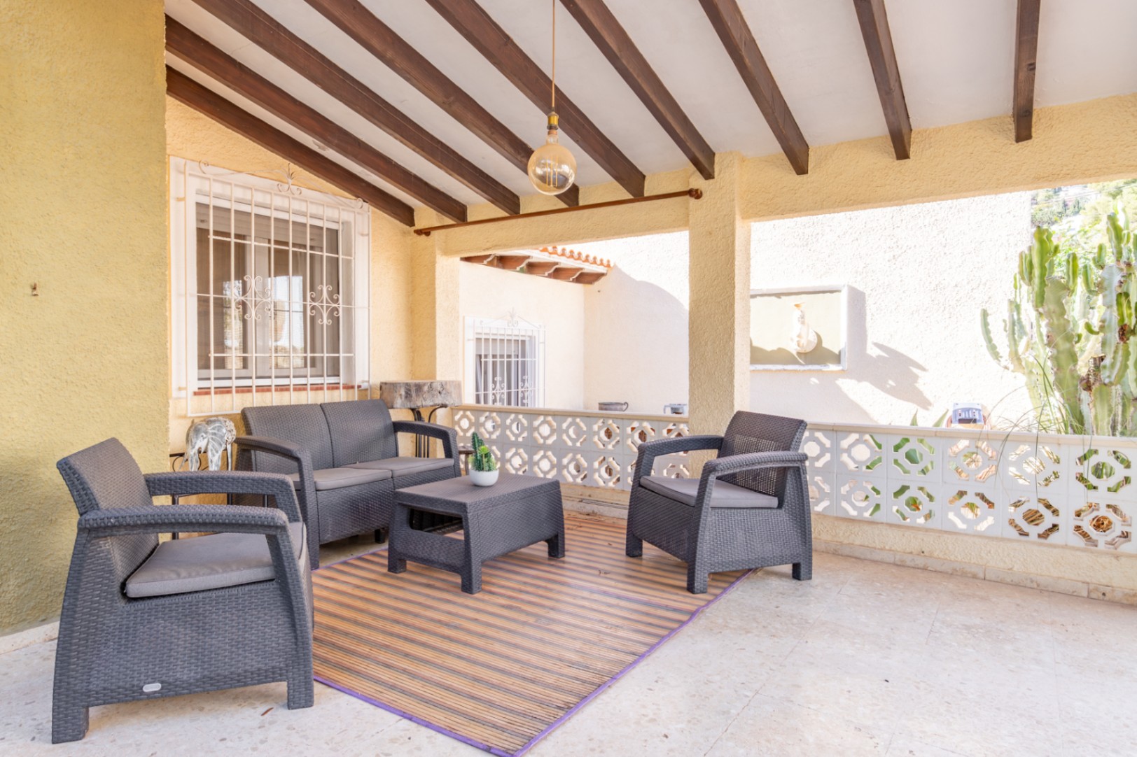 Terraza cubierta con vigas de madera vista, mobiliario de resina trenzada gris y balaustrada decorativa tradicional con vistas al jardín mediterráneo.