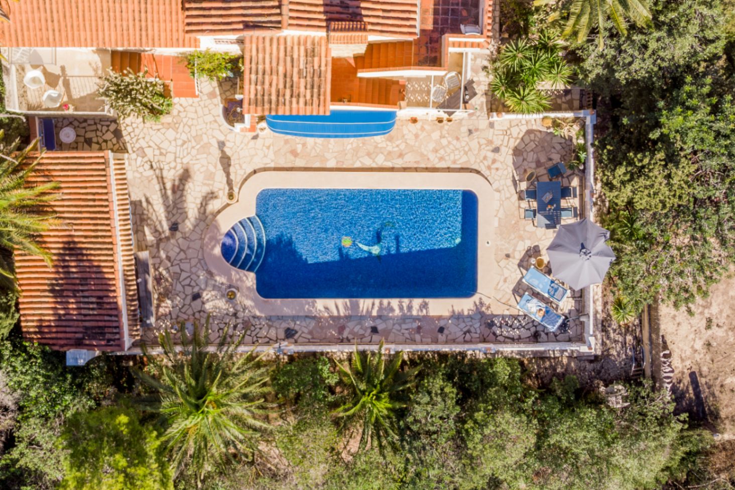 Vista aérea de la amplia terraza de piedra natural con piscina de 42 m², zona de comedor exterior y solárium rodeado de vegetación mediterránea.