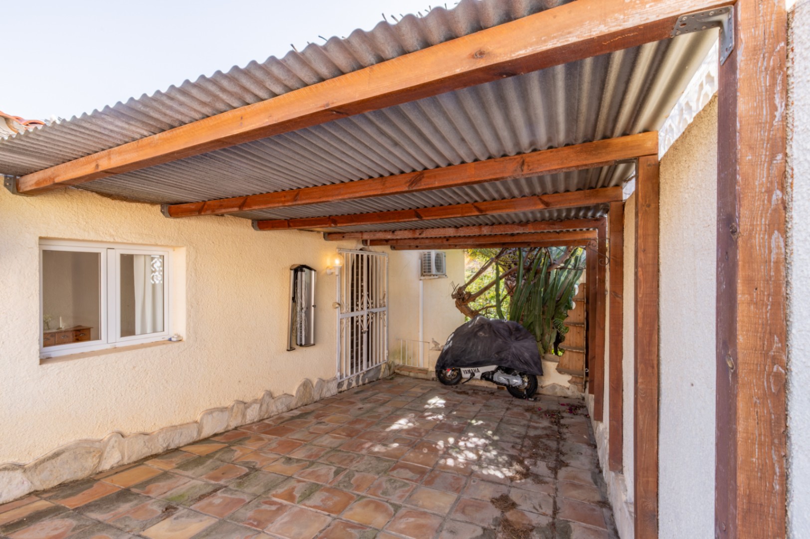 Zona de aparcamiento cubierta con estructura de vigas de madera y techo ondulado, suelo de gres rústico y acceso directo a la vivienda mediante puerta de seguridad.