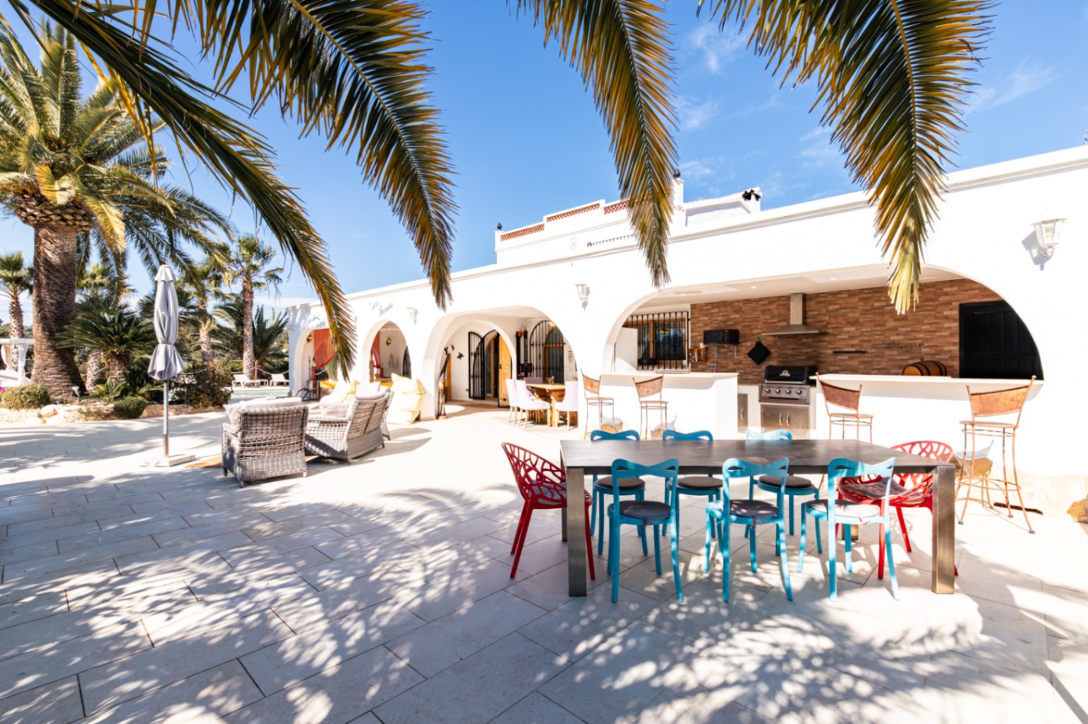 Amplia terraza exterior con naya arqueada, cocina de verano equipada con barbacoa Broil King y zona de comedor bajo palmeras maduras.