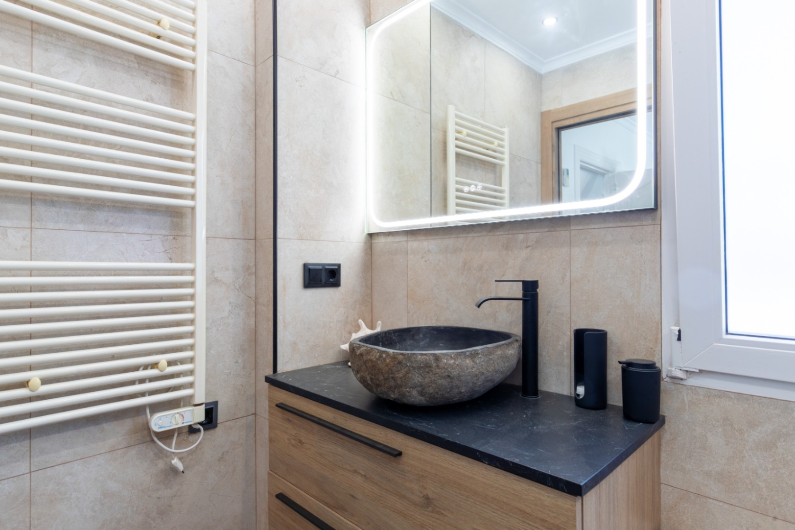 Baño moderno con lavabo de piedra natural sobre encimera negra y mueble de madera. Incluye espejo retroiluminado LED y radiador toallero blanco montado en pared.