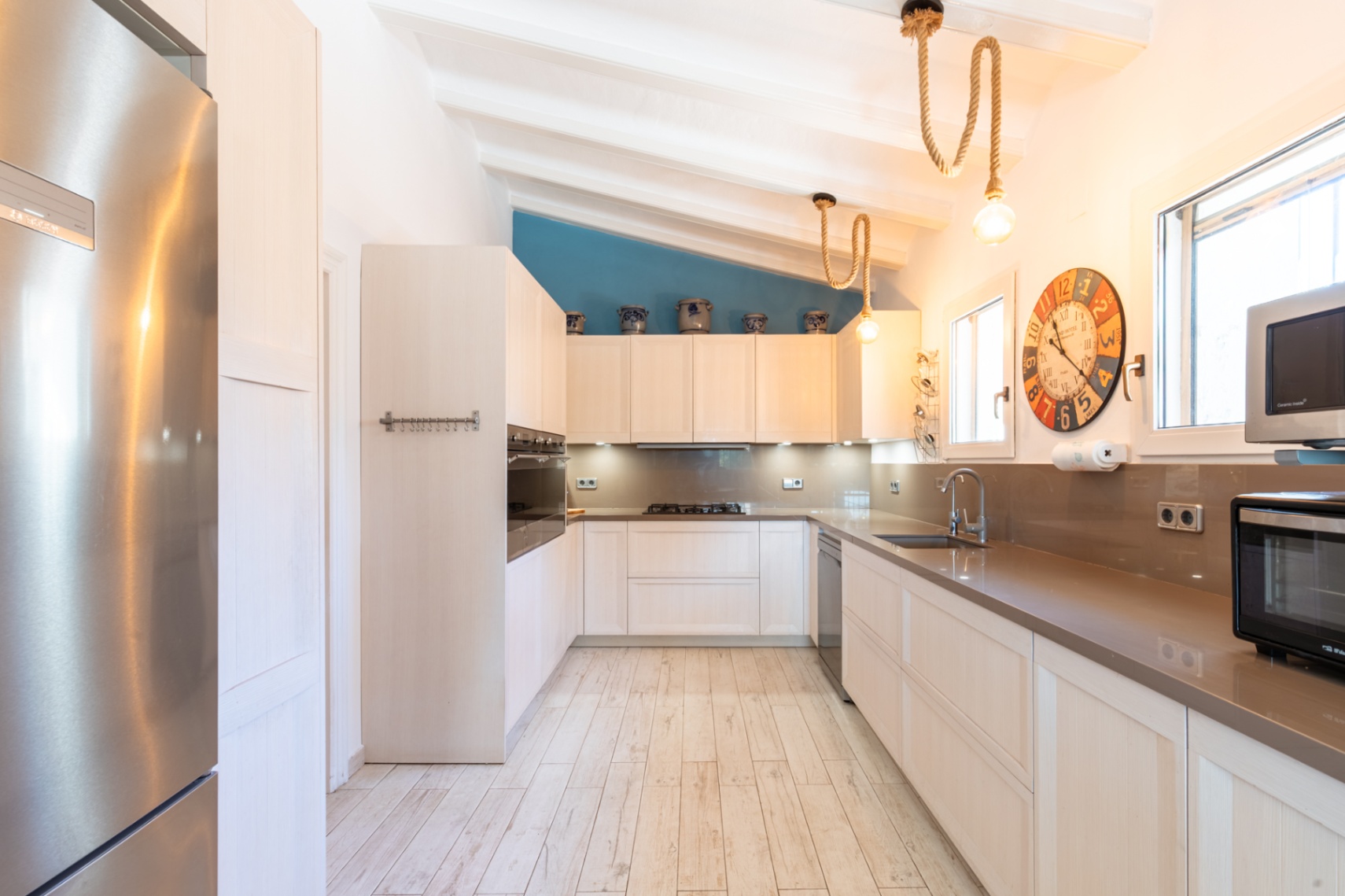 Cocina equipada con electrodomésticos de alta gama, encimeras de piedra y techo inclinado con vigas blancas. Incluye iluminación decorativa de cuerda y suelo de gres cerámico.