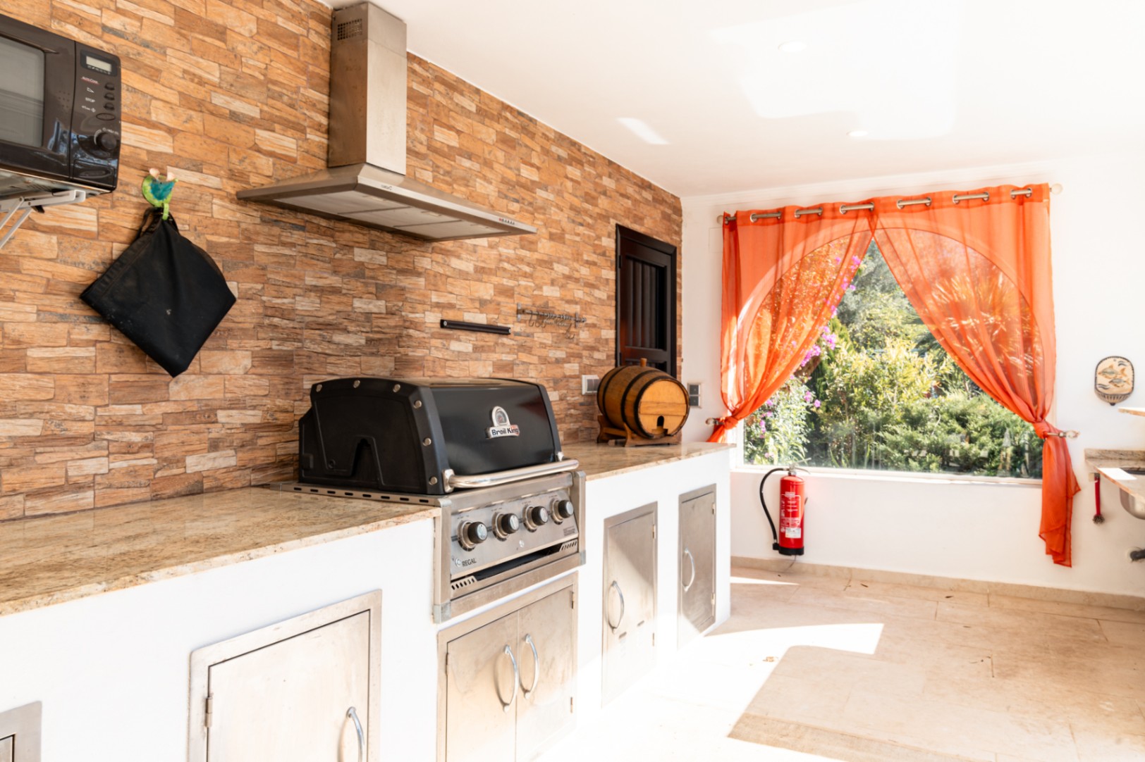 Cocina exterior equipada con barbacoa Broil King encastrada, encimera de granito y pared revestida en piedra natural bajo una campana extractora de acero inoxidable.