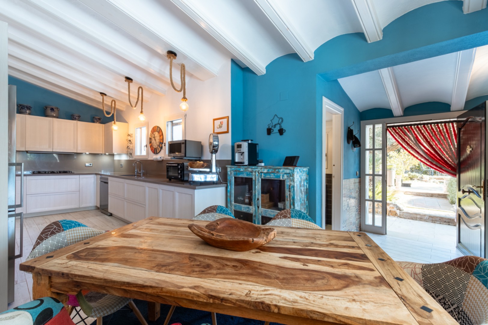 Cocina moderna con vigas blancas, paredes en azul intenso, mesa de madera rústica y lámparas de cuerda. Incluye electrodomésticos de alta gama y acceso directo al exterior.