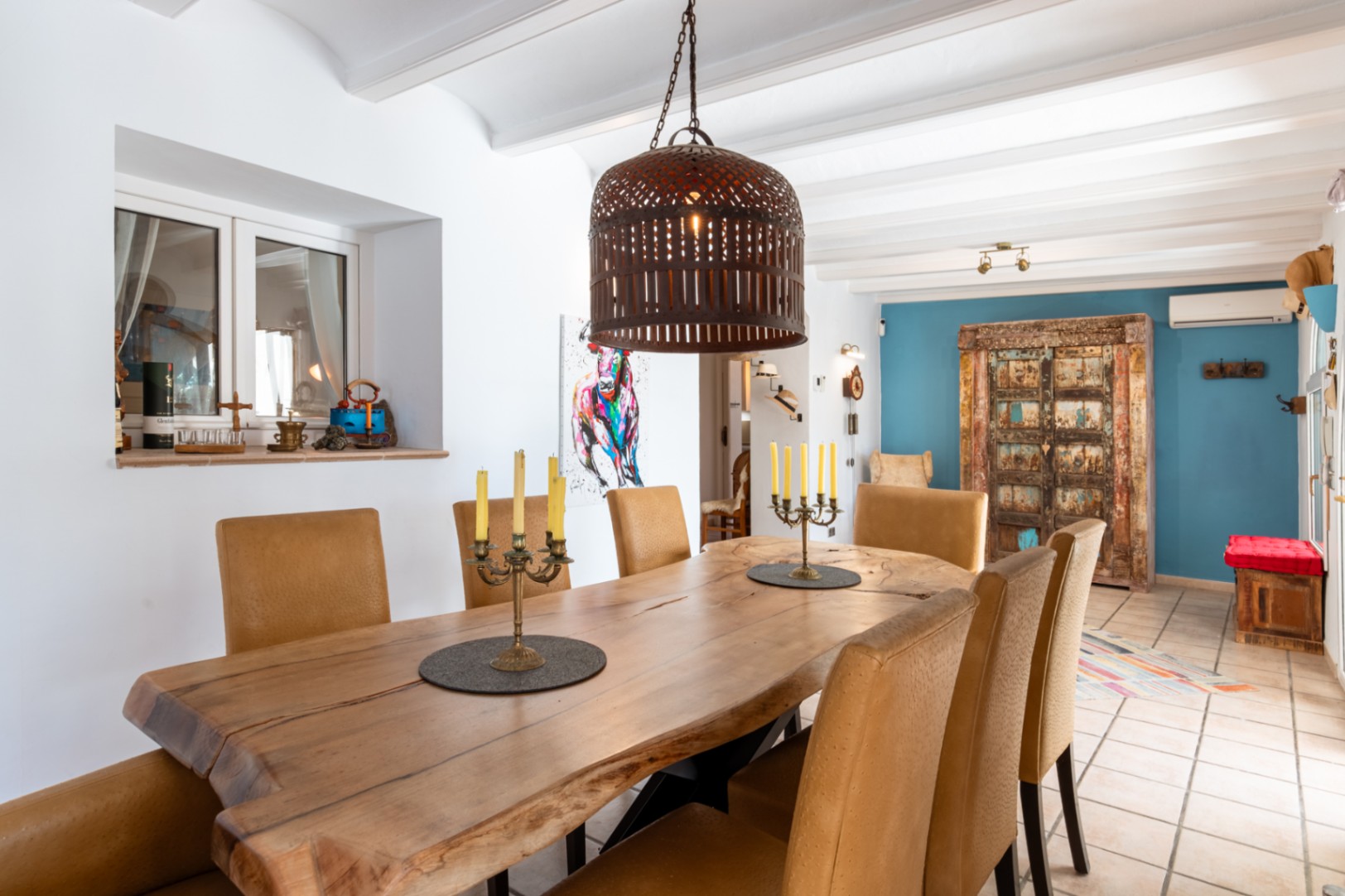 Comedor con mesa de madera maciza de borde natural, sillas tapizadas y techo de vigas blancas, complementado por una pared de acento azul y detalles rústicos.