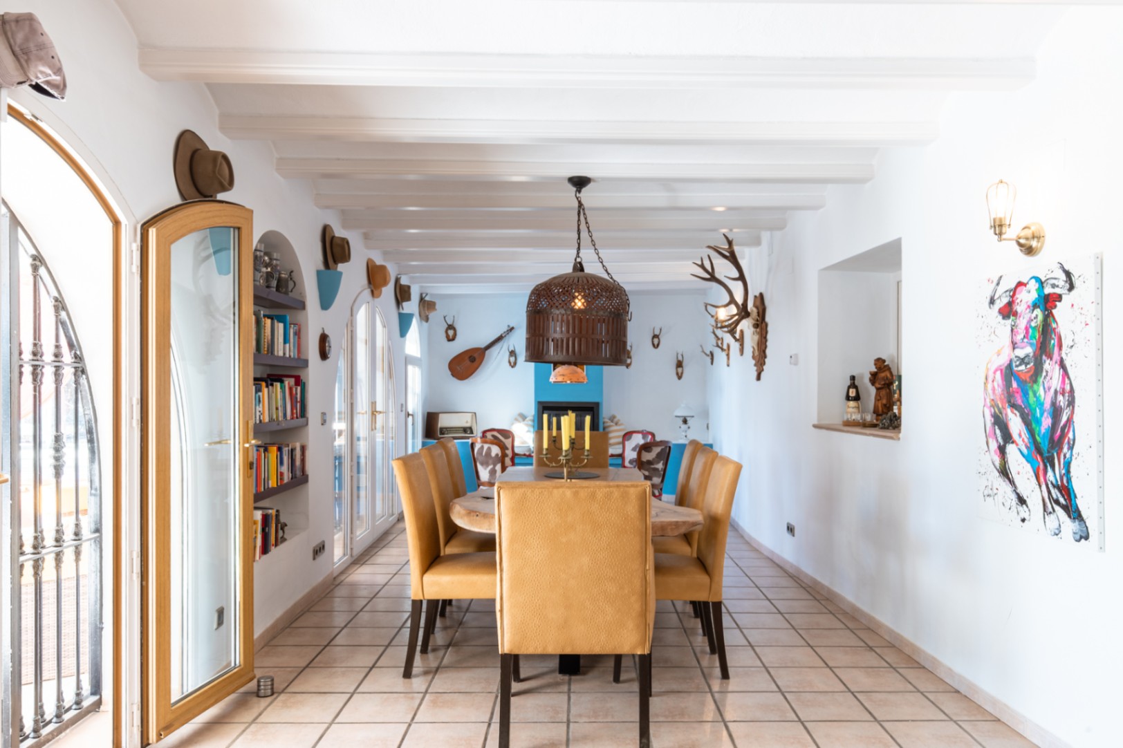 Comedor espacioso con vigas blancas expuestas, mesa de madera maciza y sillas tapizadas. La estancia cuenta con puertas de arco, estanterías integradas y una decoración rústica ecléctica.