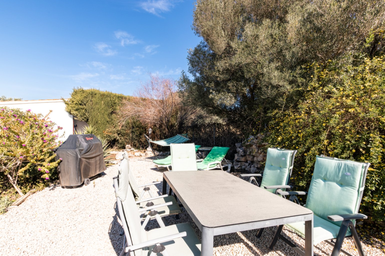 Comedor exterior sobre grava con mesa rectangular, sillas acolchadas, barbacoa protegida y hamaca, rodeado de vegetación autóctona bajo un cielo despejado.