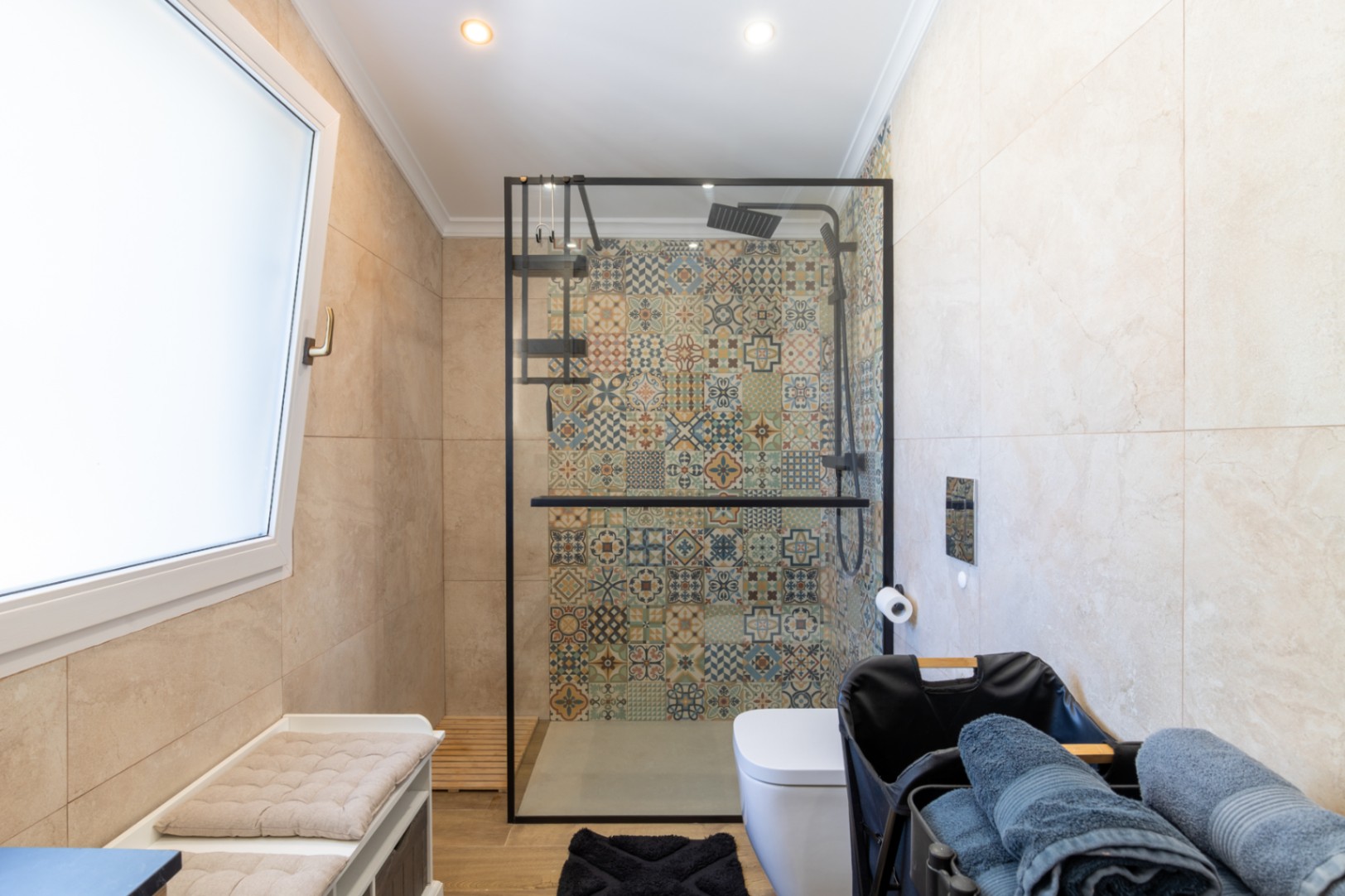 Cuarto de baño moderno con ducha a ras de suelo, destacando una pared de azulejos con motivos geométricos, mampara de perfilería negra y suelos de gres efecto madera.