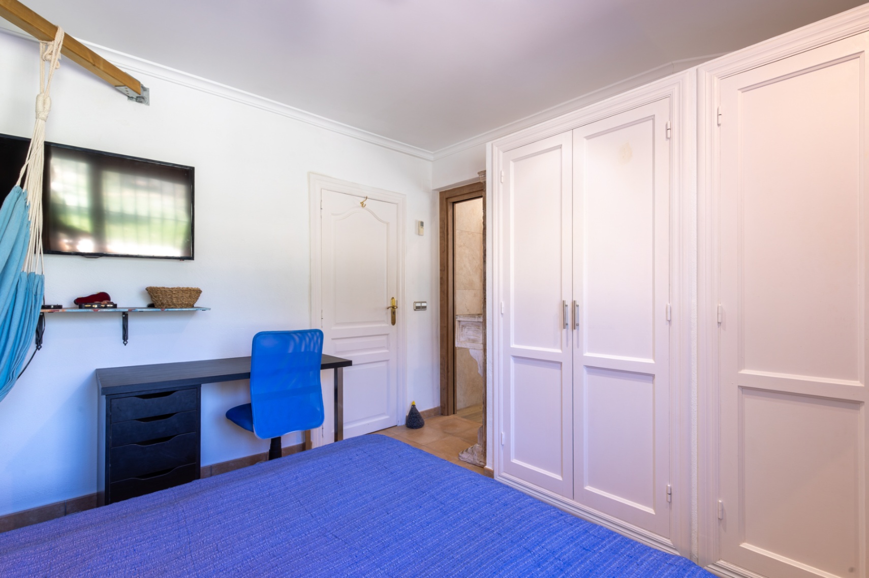 Dormitorio con armarios empotrados blancos, zona de escritorio con silla azul y acceso directo a un baño privado con acabados en piedra natural.