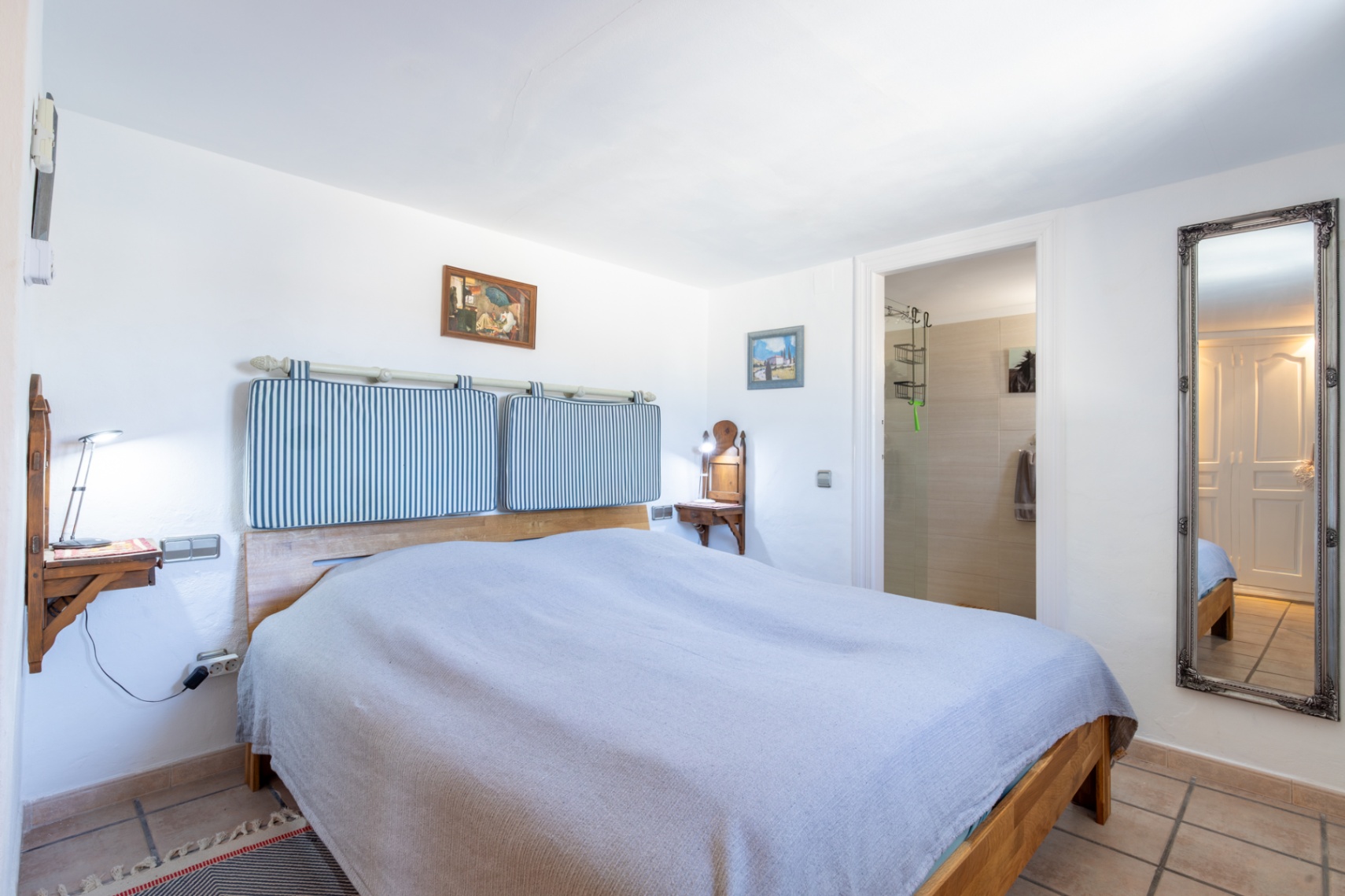 Dormitorio con cama de madera, cabecero de cojines a rayas y mesitas de noche integradas. Incluye acceso directo a baño en suite y un gran espejo de pared con marco decorativo.