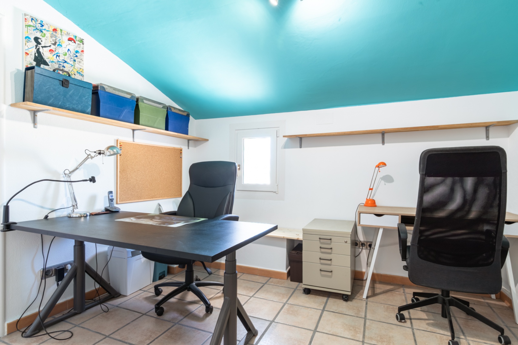 Oficina luminosa con techo inclinado en color turquesa, equipada con dos puestos de trabajo, estanterías de madera, suelo cerámico y luz natural.