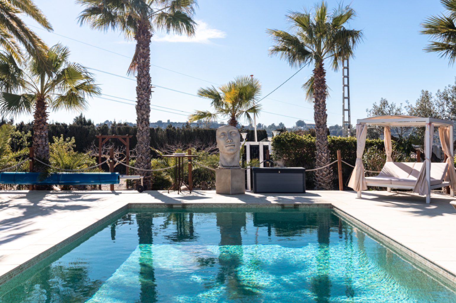 Piscina de 9,5m x 5m rodeada de una terraza soleada con palmeras, una escultura decorativa, cama balinesa y sistema de cubierta enrollable para el mantenimiento del agua.