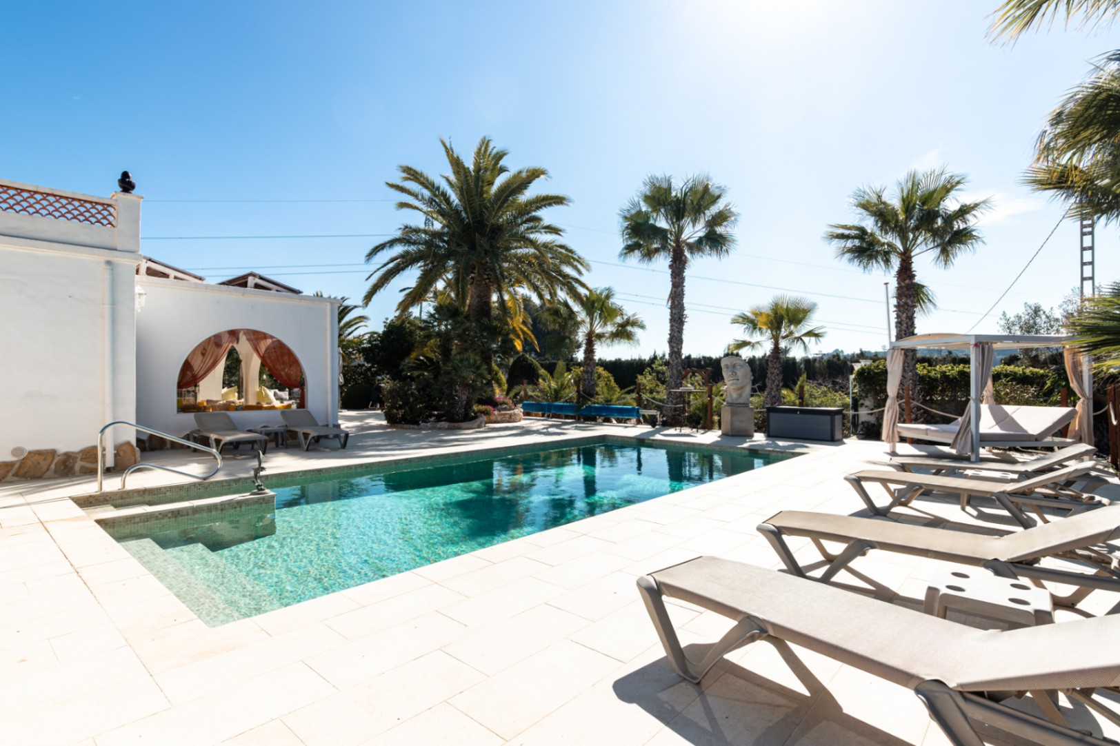 Piscina de 9,5x5m con terraza de piedra clara, zona de tumbonas, cama balinesa y jardín mediterráneo con palmeras bajo el sol.