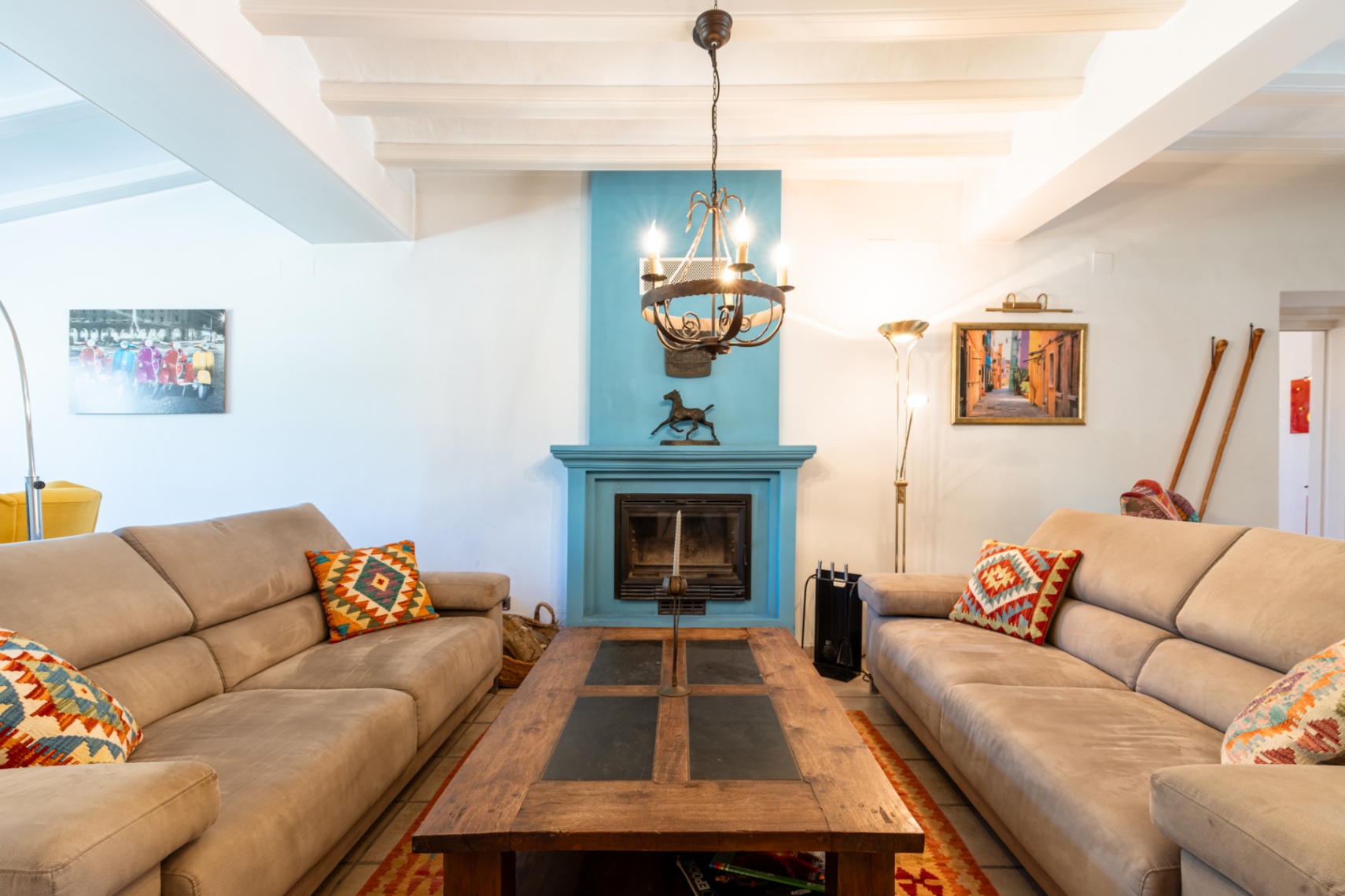 Salón luminoso con vigas de madera blancas, chimenea central en azul vibrante y sofás enfrentados alrededor de una mesa de centro de madera maciza y pizarra.