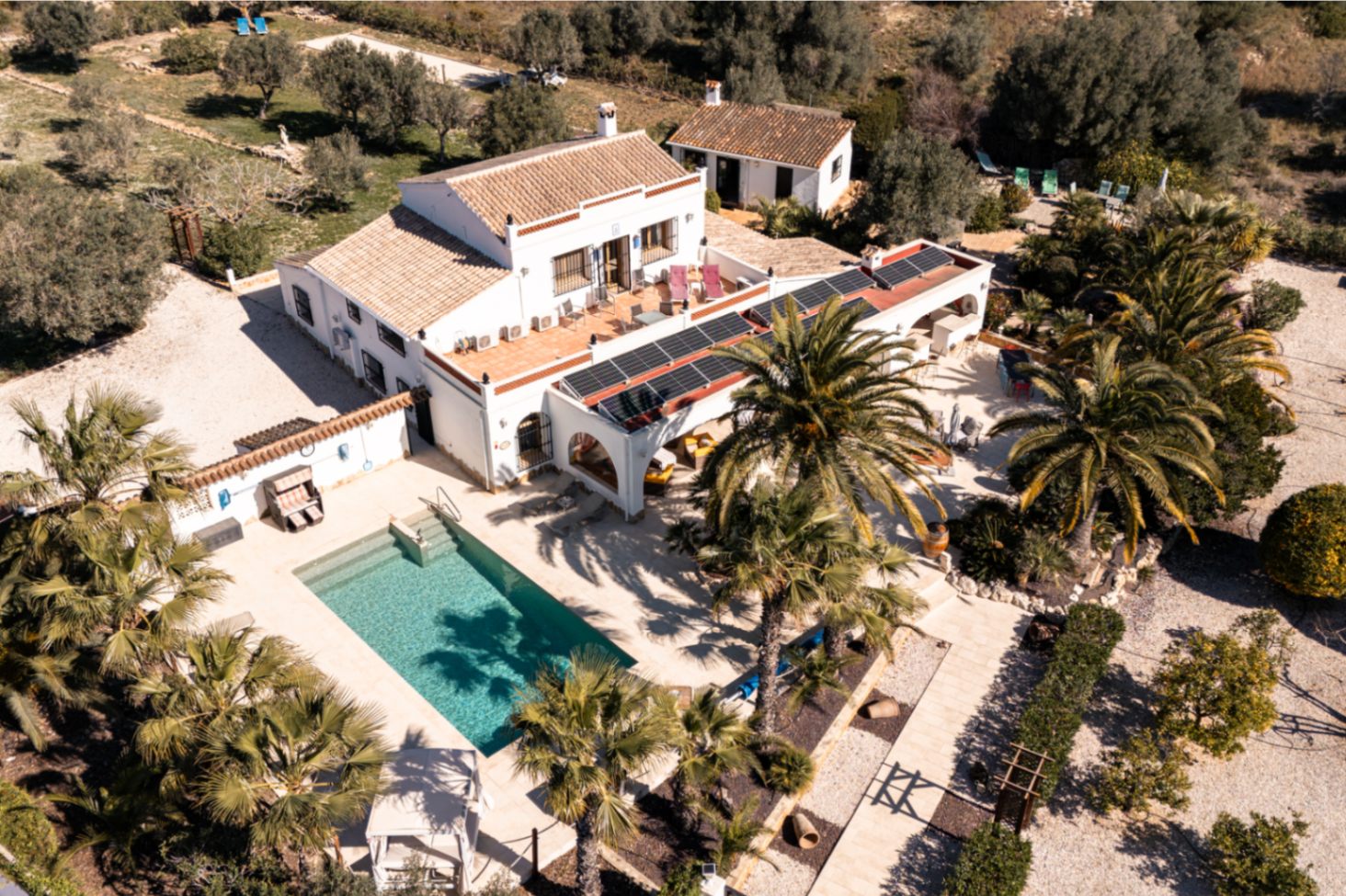 Vista aérea de la finca con piscina rectangular, amplias terrazas y paneles solares integrados en la arquitectura tradicional rodeada de vegetación mediterránea.