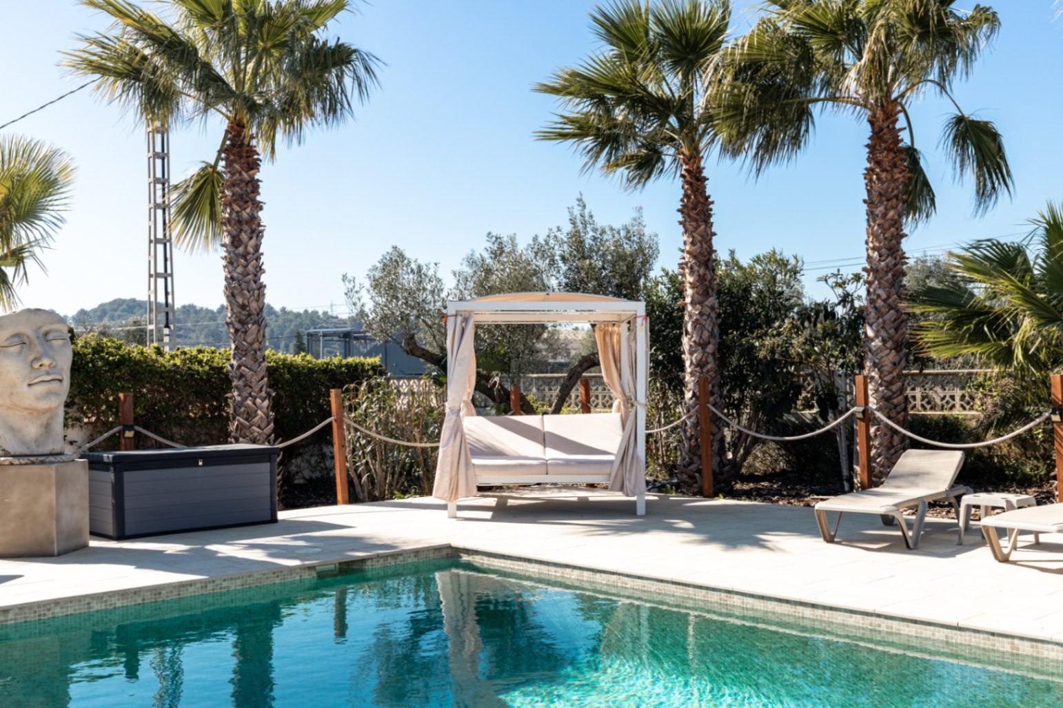 Zona de piscina con solárium de piedra natural, cama balinesa con cortinas, tumbonas y vegetación mediterránea con palmeras bajo un cielo despejado.
