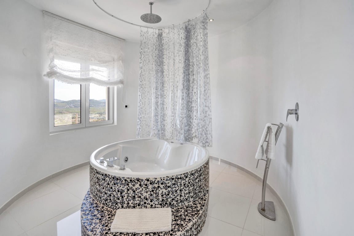 Baño de diseño con bañera circular de hidromasaje decorada con mosaicos, ducha de efecto lluvia integrada y ventanal con vistas a la montaña.