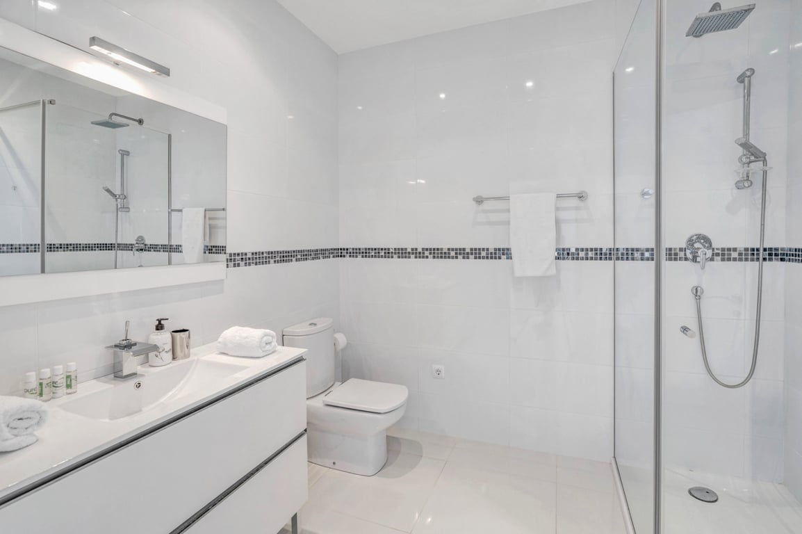 Baño moderno con azulejos blancos y cenefa de mosaico, mueble de lavabo suspendido, inodoro y ducha de obra con mampara de cristal y grifería cromada.