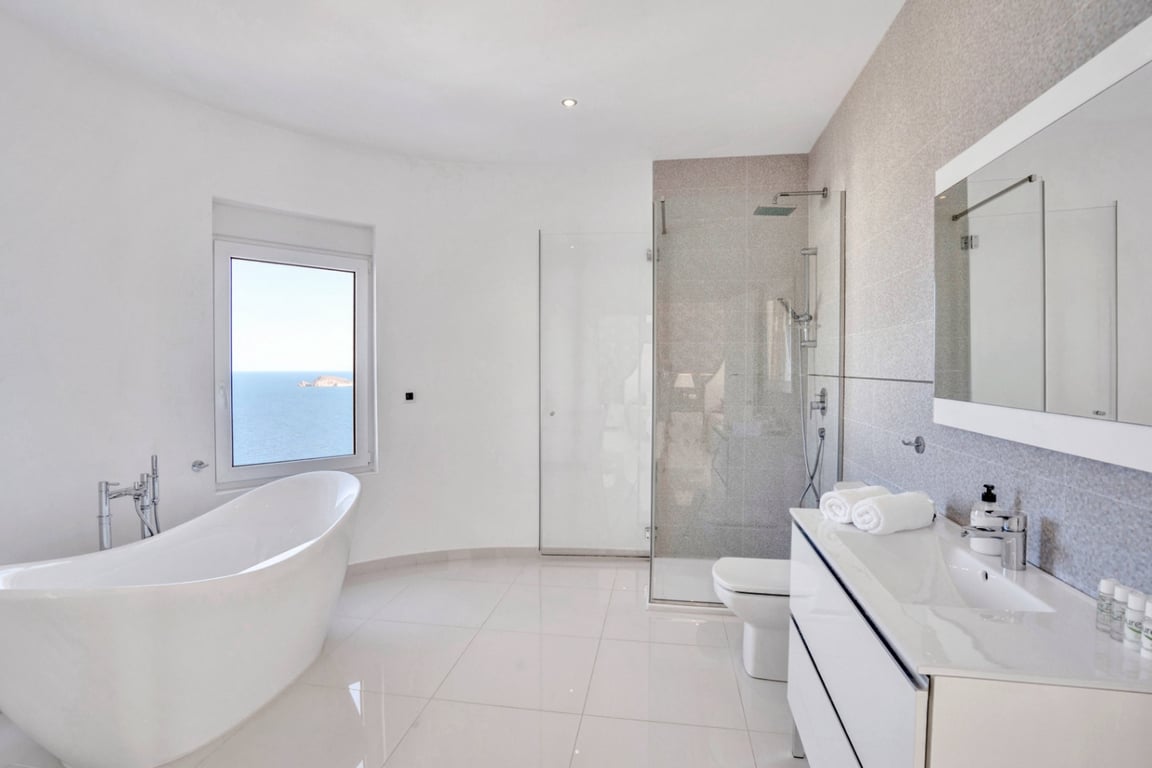 Baño moderno con bañera exenta junto a ventana con vistas al mar, ducha de obra acristalada y mueble de lavabo blanco con espejo de gran formato.
