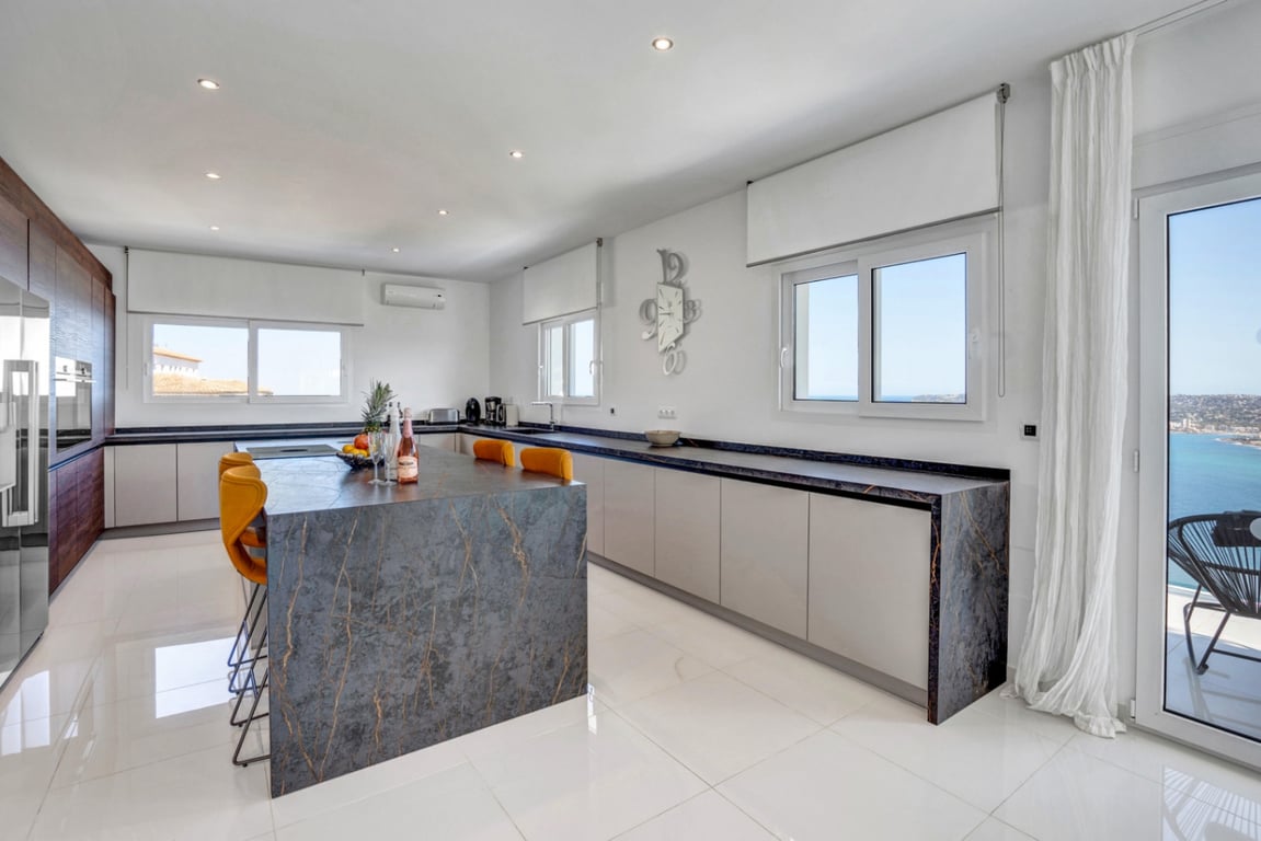 Cocina de diseño con isla central de piedra veteada, taburetes mostaza y armarios minimalistas. El espacio cuenta con mucha luz natural y vistas directas al mar Mediterráneo.