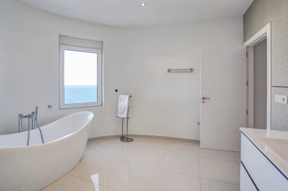 Cuarto de baño moderno con bañera exenta de diseño, suelos cerámicos claros y ventana con vistas directas al mar Mediterráneo.